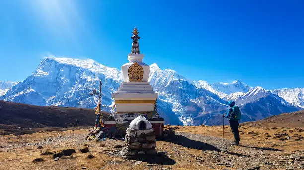 A man standing next to a stupa with snow caped Annapurna chain in the back, Annapurna Circuit Trek, Himalayas, Nepal. High mountains around. Some prayer's flag next to it. Serenity and calmness.