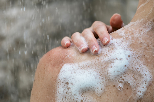 Cropped view of young adult girl taking shower, standing in bathroom with copy space and flowing water on background, holding hand on shoulder with soap foam