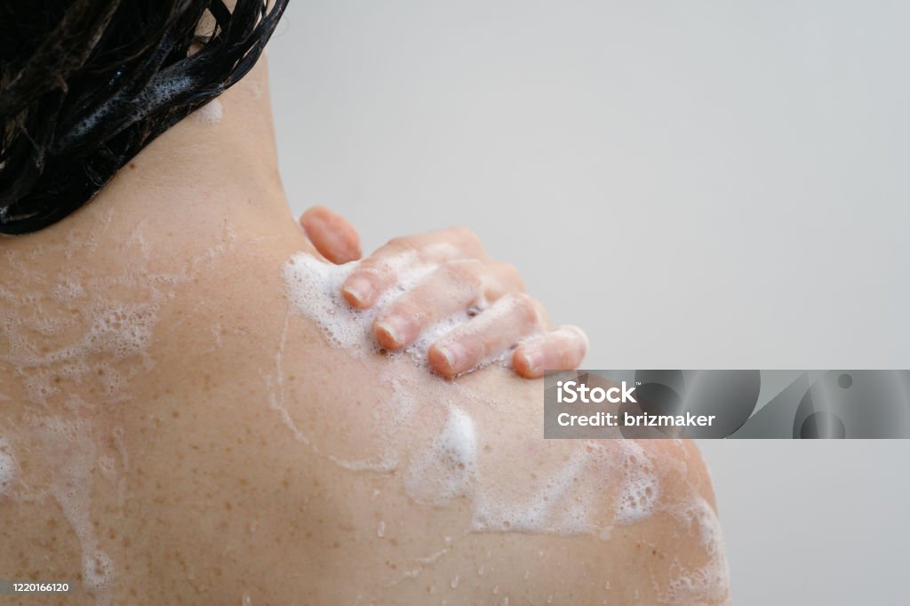 Young adult girl taking shower, standing in bathroom Cropped back view of young adult girl taking shower, standing in bathroom with copy space on background, holding hand on shoulder with soap foam Shower Stock Photo