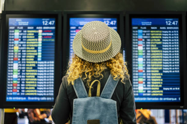 Travel people in airport or train station concept - view from rear of a blonde curly female woman with blue backpack looking and checking time departures or arrivals on the displays screen Travel people in airport or train station concept - view from rear of a blonde curly female woman with blue backpack looking and checking time departures or arrivals on the displays screen delayed sign photos stock pictures, royalty-free photos & images
