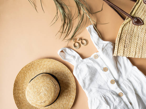 Women's summer straw hat, wicker bag, white sundress, sunglasses and jewelry on a beige background. Copy space. Top view stock photo