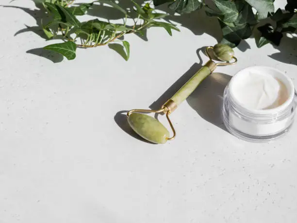 Photo of Green Gua sha massage jade roller and face cream in white container on white background.