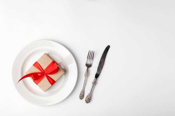 istock Anniversary, birthsday festive romantic table setting on white background. Kraft gift box with red ribbon in ceramic plate, silverware, vintage fork, knife. Place for text, copyspace, top view 1220173108
