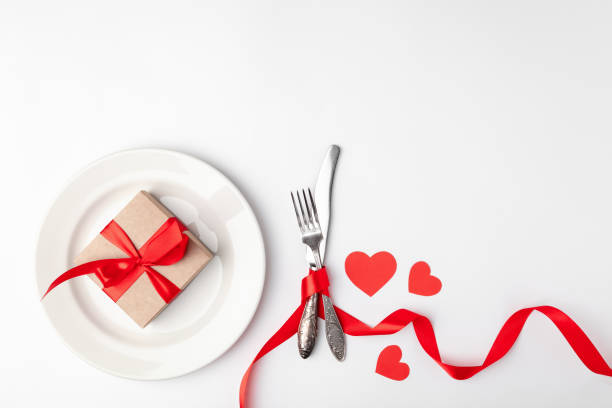 istock Romantic table setting on white background. Valentines day card template. Kraft gift box with red ribbon in plate, silverware. Concept anniversary, mother day, birthday greeting, copyspace, top view 1220173222