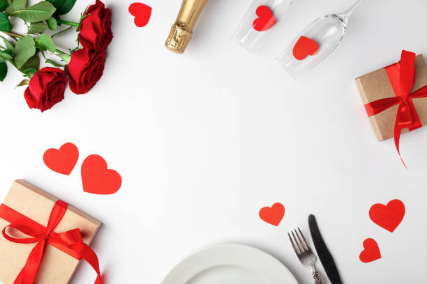 istock Valentines day dinner with table place setting, red tape, kraft gift box, glass for champagne, champagne bottle, roses, hearts, silverware on white background. Copyspace. Space for text. Top view 1220173240