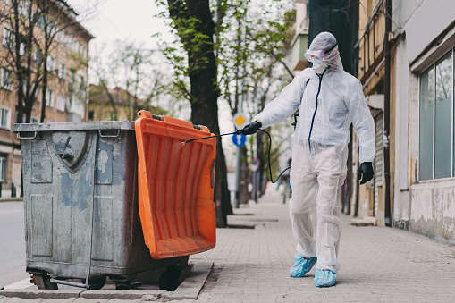 Man in protective suit spraying with disinfectant garbage cans and pavements to prevent further spread of the coronavirus