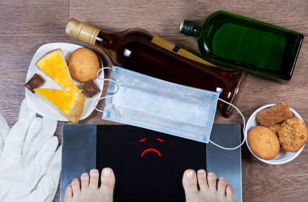 woman checks her weight after quarantine. sad smile on balance screen. bottles of alcohol, sweet food, face mask and gloves on the floor. - dieting overweight weight scale healthcare and medicine imagens e fotografias de stock