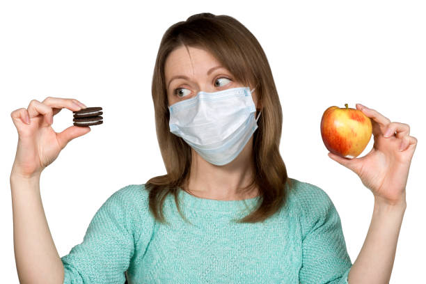 Young woman in face mask is choosing between sweets and apple. Concept of healthy eating during quarantine and self-isolation. stock photo