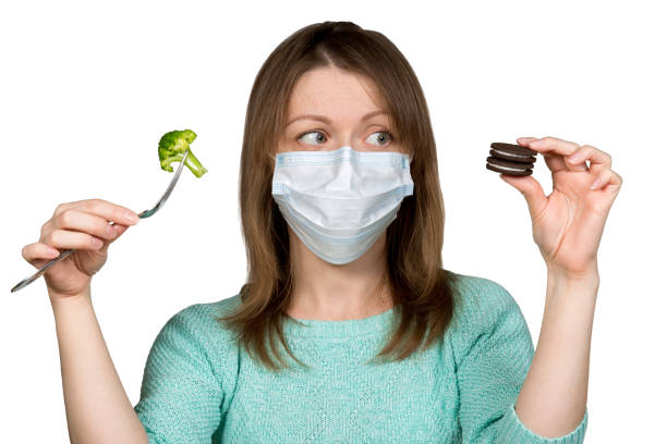 Young woman in face mask is choosing between sweets and brocoli. Concept of healthy eating during quarantine and self-isolation. stock photo