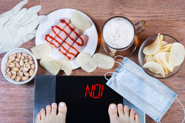 Girl checks her weight after quarantine. Digital scales with word no on screen. Beer, plates with junk food, face mask and gloves on the floor. stock photo