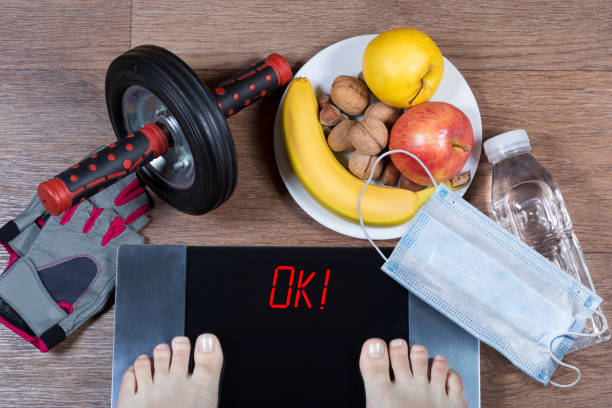 Girl checks her weight after quarantine. Digital scales with word ok surrounded by sport accessories, healthy food, water bottle and face mask. stock photo