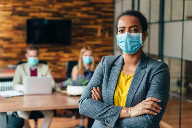 Businesspeople wearing masks in the office for safety during COVID-19 pandemic, mixed race businesswoman looking at camera
