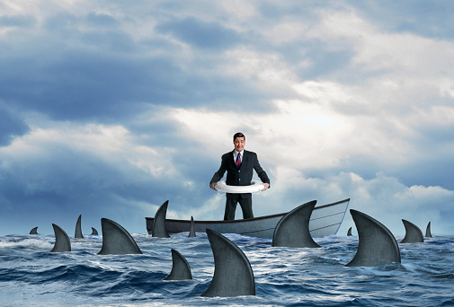 A fearful businessman wearing a life preserver around his waist as he stands in a wooden dinghy that is surrounded by a school of sharks.