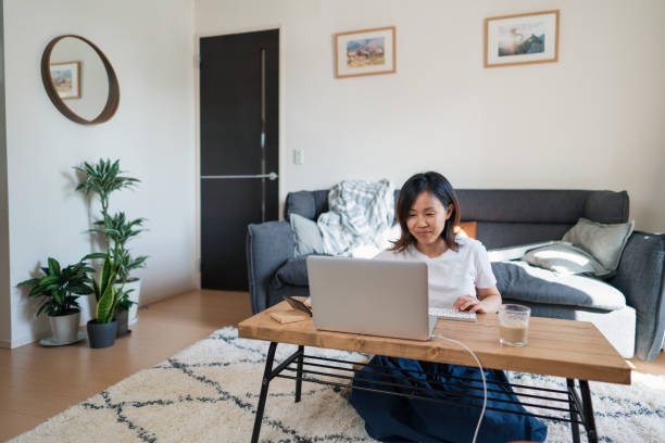 woman working on her laptop at home in her living room - japanese culture asian ethnicity friendship computer imagens e fotografias de stock