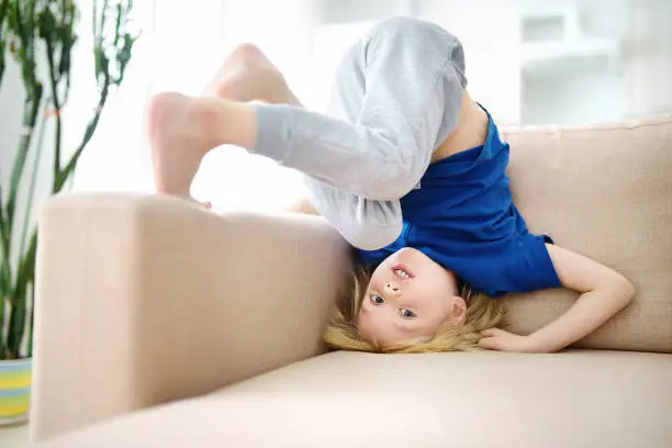 Little boy is standing on own head on sofa at home while pandemic. Bored lonely child. Difficulties of family with preschool children during quarantine. Stay at home.