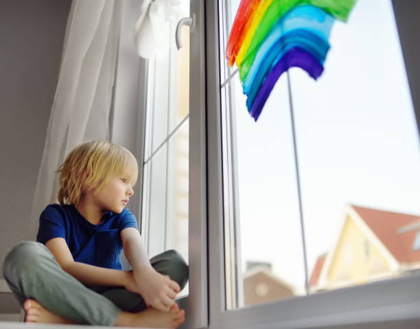 istock Little boy sitting at window with drawing rainbow while coronavirus quarantine. Rainbow sign is symbol of hope, means that everything will be OK. 1220177941