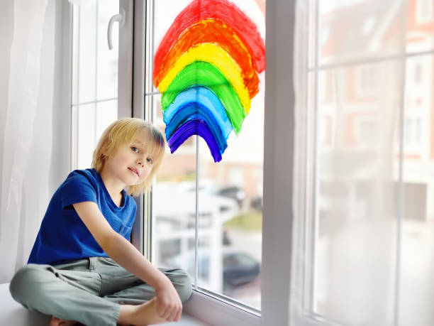 istock Little boy sitting at window with drawing rainbow while coronavirus quarantine. Rainbow sign is symbol of hope, means that everything will be OK. 1220178019