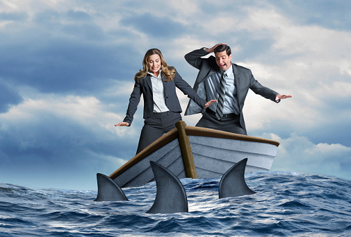 A fearful businessman and businesswoman look down in fear as they stand in a wooden dinghy that is surrounded by a school of sharks.