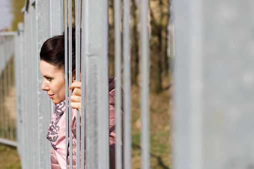A girl looks through the bars of a metal fence. Dressed in a spring jacket, a scarf around her neck.