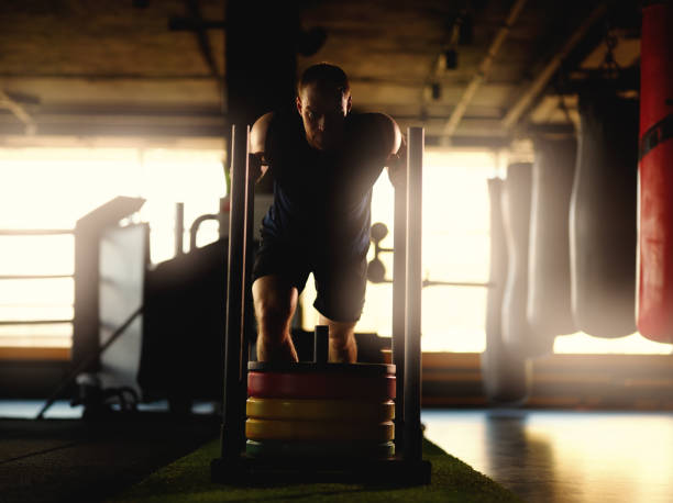 istock Full length backlit shot of athletic middle aged man pulling weight sled with plates on artificial lawn grass during workoutin gym 1220199595