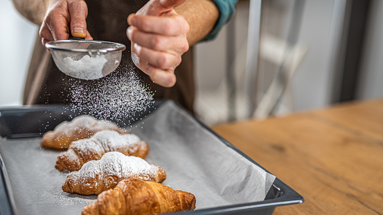 Close-up of chef's hand sprinkling powdered sugar on croissant with sieve in kitchen.