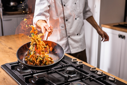 Close-up of chef's hand tossing and flaming vegetables in pan.