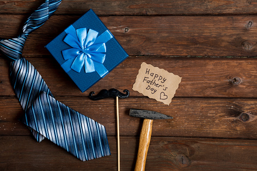 Happy Fathers Day gift box with tie, hammer, blue box and mustache on a rustic wood background. Space for text.