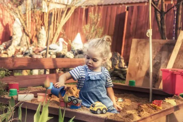 Happy beautiful child girl playing alone outdoors on playground with toy cars in sandbox in countryside concept for childhood lifestyle