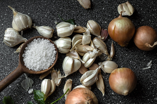 On the surface are garlic cloves, onions and salt in a wooden spoon.