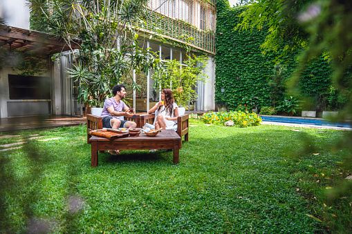 Mid distance view of young mixed race and Hispanic couple relaxing outdoors in backyard with breakfast and weekend leisure.