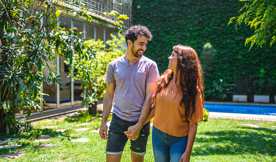 Close-up of smiling Hispanic and mixed race couple face to face and walking in sunny backyard holding hands.