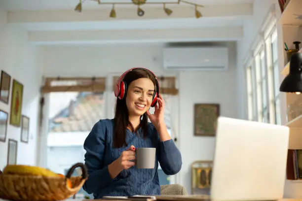 Attractive young woman teacher, teaching by holding an online class with her students from home using her laptop. Having a headphones on and smiling. Relaxed atmosphere and cozy ambient for the improvement of productivity.