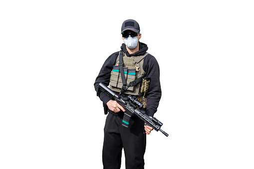 Army Man wearing Tactical Uniform and holding Machine Gun in Hands and Aiming. Isolated on White Background. Black Clothes and Hoody. White Mask against Coronavirus, disease or virus