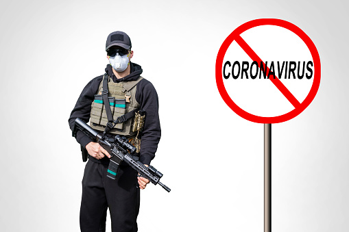 Coronavirus Ban Sign. Army Man wearing Tactical Uniform and holding Machine Gun in Hands and Aiming. Isolated on White Background. Black Clothes and Hoody