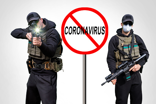 Coronavirus Ban Sign. Army Man wearing Tactical Uniform and holding Machine Gun in Hands and Aiming. Isolated on White Background. Black Clothes and Hoody