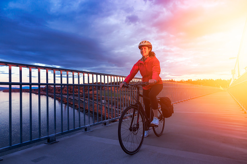 Caucasian Woman riding a Bicycle on a path at Port Mann Bridge during vibrant Sunset. Taken in Surrey, Vancouver, British Columbia, Canada.