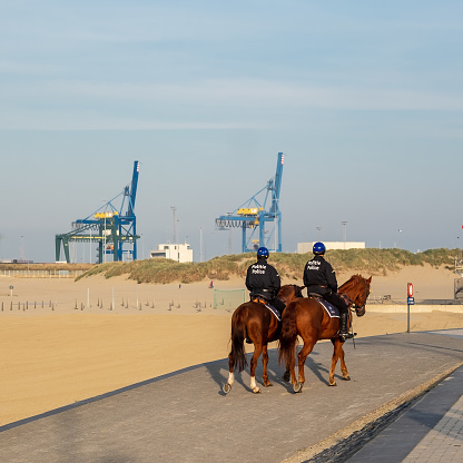 Zeebrugge, Belgium - 31 October 2019: Two Belgian police agents on horses on the beach of Zeebrugge, with cargo cranes of the port in the background
