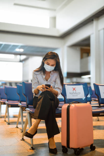 jovem turista asiática com máscara usando telefone e sentada com distância no aeroporto - airport lounge airport one person bag - fotografias e filmes do acervo