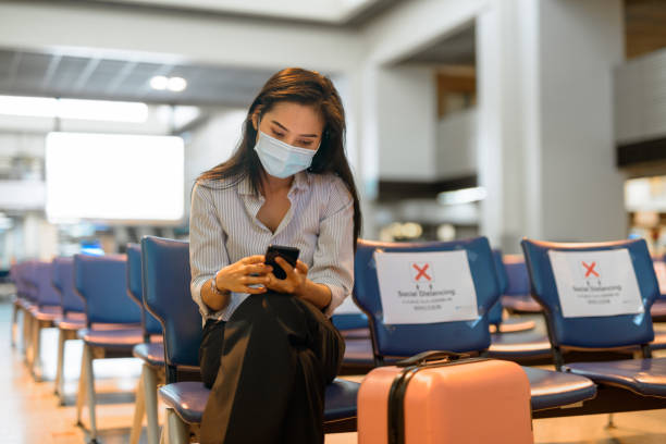 jovem turista asiática com máscara usando telefone e sentada com distância no aeroporto - airport lounge airport one person bag - fotografias e filmes do acervo