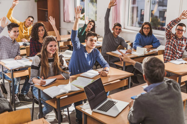 Large group of happy high school students raising their hands on a class. Large group of happy students raising their hands to answer teacher's question on a class at school. Focus is on boy in blue sweater. teenage high school girl raising hand during class stock pictures, royalty-free photos & images