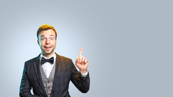 Studio shot of a young man pointing up with one index finger. He is smiling excitedly and wearing a bow tie. Neutral background with copy space.