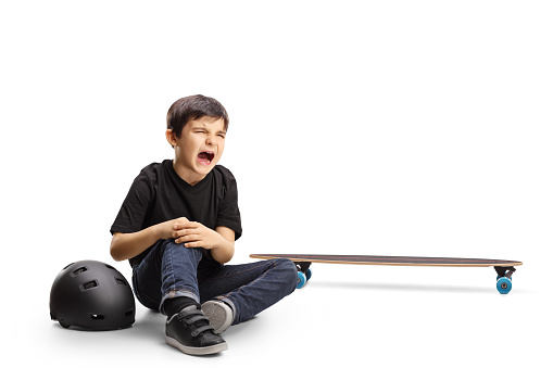 Child sitting on the floor crying and holding knee hurt from a skateboard isolated on white background
