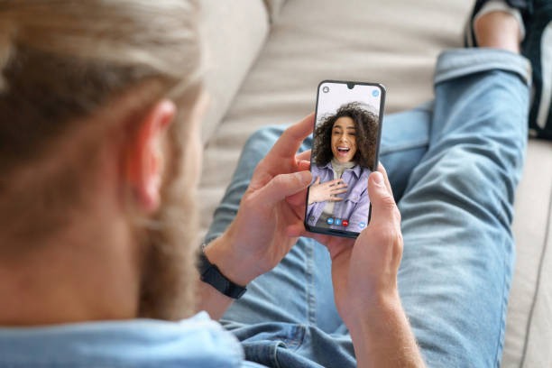 young man sitting on sofa holding smartphone communicating with african girl friend on mobile screen, making video call using cell phone mobile social media dating app. video call concept. over shoulder closeup view - internet dating imagens e fotografias de stock
