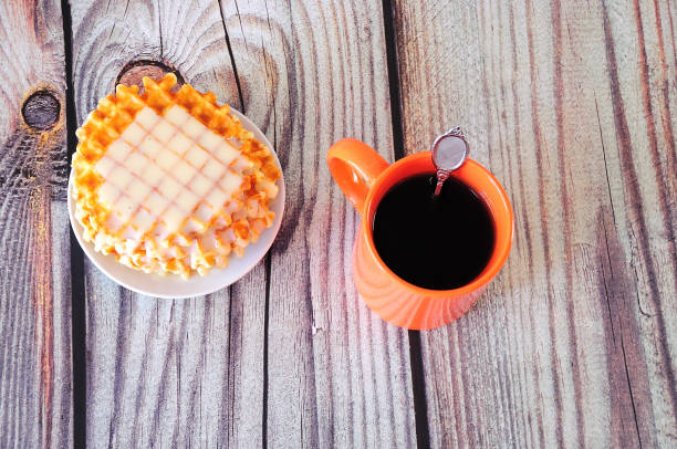 An orange ceramic mug of hot coffee and a plate with Parisian waffles and creamy syrup.