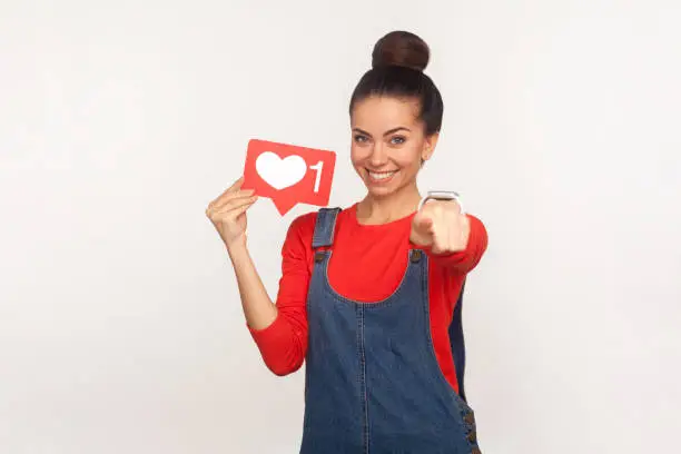 Photo of Hey you, like and follow interesting content! Portrait of positive stylish girl in denim overalls holding social media heart icon