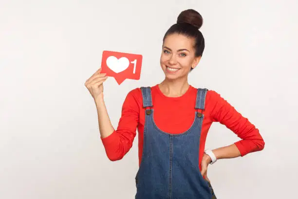 Photo of Like and follow interesting content! Portrait of happy stylish trendy girl in denim overalls holding social media heart icon