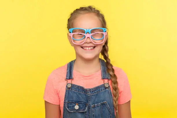 Photo of Eyewear fashion. Portrait of funny little girl in denim overalls wearing two stylish eyeglasses and smiling joyfully