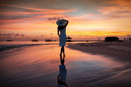 Tropical travel concept with a woman with a sunhat standing on a paradise beach and enjoying the fiery sunset during her vacation time