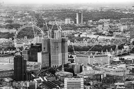 This aerial black and white photograph presents a timeless view over London's iconic skyline. Dominating the scene is the majestic silhouette of the Houses of Parliament and the iconic clock tower, Big Ben, standing as sentinels over the River Thames. The river's calm waters reflect the grandeur of these historical structures, while the sprawling urban landscape stretches into the horizon, peppered with both modern and ancient buildings. The overcast sky casts a diffuse light, lending the city a soft, ethereal quality and highlighting the intricate gothic architecture. This image encapsulates the enduring legacy and the unchanging face of London amidst the ever-evolving tapestry of city life, inviting the viewer to ponder the stories and history woven into the fabric of this grand metropolis.