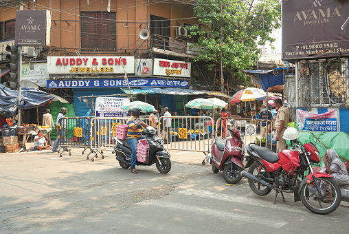 Road partially blocked by kolkata police guardrails, to block entry of cars & unwanted people in marketplace. A motorbike rider is seen waiting there, while a kolkata police guard in keeping an eye on him.\n\nPhoto taken at Jadubabur Bazar, Bhowanipore Kolkata, 4/20/2020.